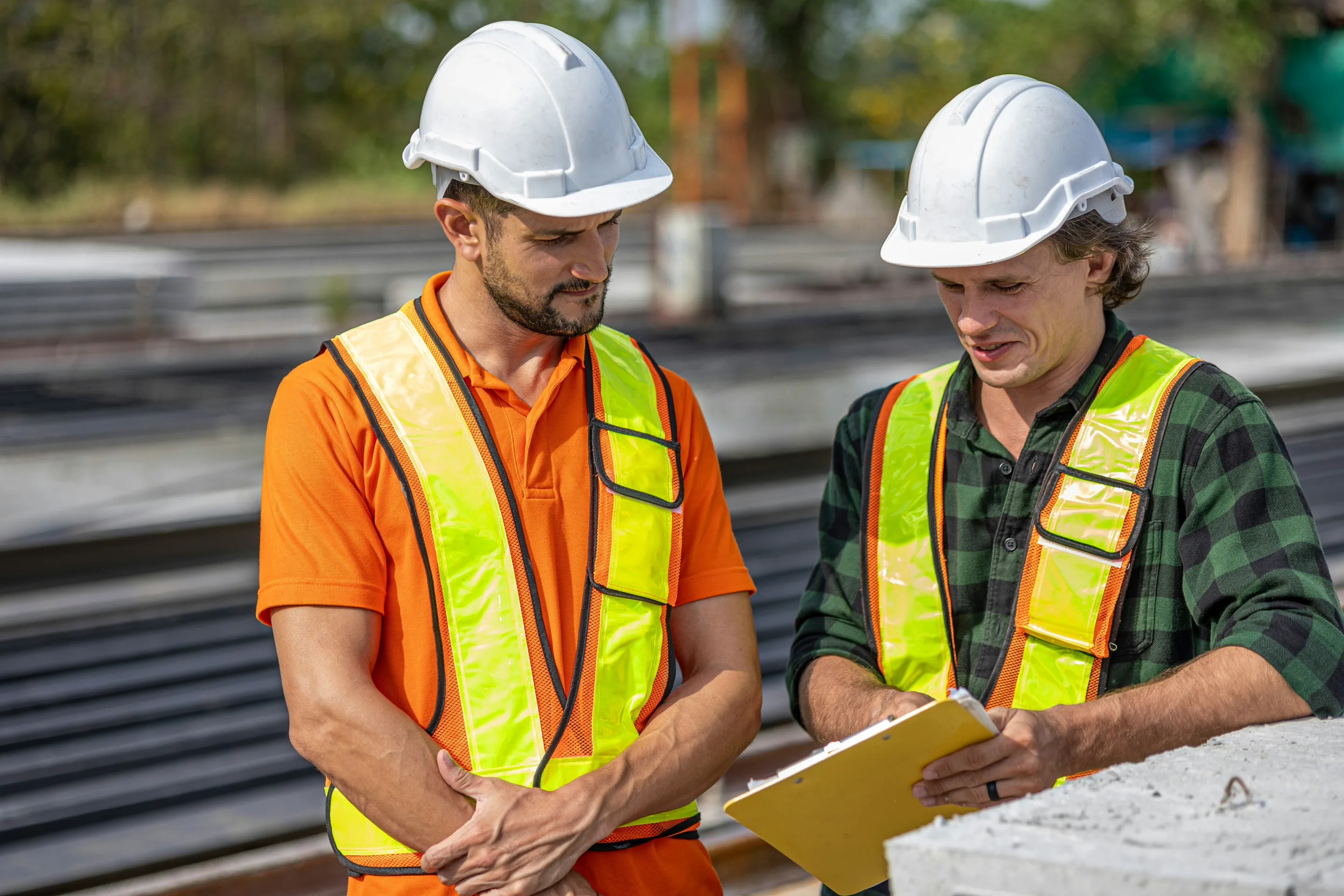 Two construction workers in hard hats and high-visibility vests reviewing a clipboard at an active worksite.